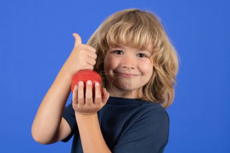 Foto de Alimentación saludable. Un chico con pulgares arriba comiendo manzana. Retrato de niño lindo niño de pie aislado sobre fondo azul - Imagen libre de derechos
