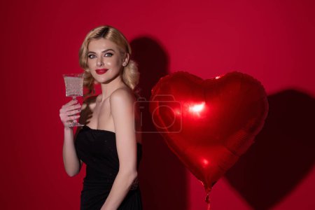Foto de Young beautiful sexy woman drink champagne. Beautiful girl with heart shape air balloon on studio isolated background. Woman on Valentines Day. Heart of love. Female model hold red balloons heart - Imagen libre de derechos