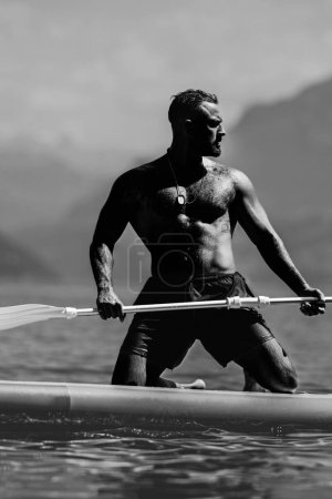 Kräftiger muskulöser Mann mit Paddelbrett. Mann paddelt auf Paddelbrett. Muskelkräftiger hispanischer Mann auf dem Surfbrett. SUP-Surfen im Sommerurlaub am Alpensee in der Schweiz