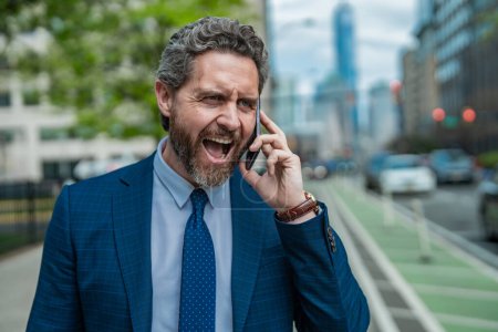 Anger Businessman with phone outdoor. Angry Business talk. Anger Man walk down street talking on the phone. Stressed Man in suit screaming on the phone. Angry man shouting at his smartphone