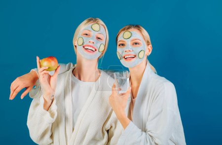 Two young happy women with facial mask and cucumber slices at face go healthy lifestyle. Girls holding glass of water and apple in hand. Natural cosmetics and health concept
