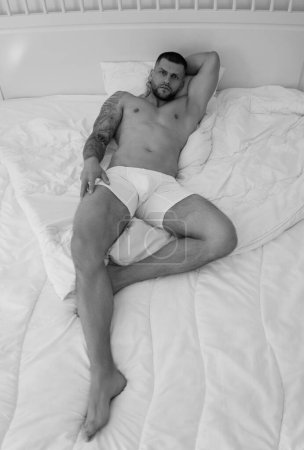 Photo for Seductive gay. Muscular body of sexy man in bed. Strong brutal guy. Sexy male naked torso. Nude muscular body man with tattoo lay in the bed. Muscular guy laying on bed - Royalty Free Image