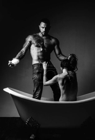 Brutal bearded tattooed man with sexy bare woman lying in bath. Handsome muscular man holds champagne cooler and bottle. Beautiful girl opens mans fly before sex. Couple foreplay