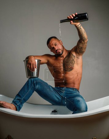 Strong muscular tattoed man holding champagne bottle and posing in bathroom. Handsome bearded shirtless man in jeans with sexy body in bathroom. Sexual macho man in bath