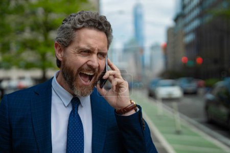 Anger Businessman with phone outdoor. Angry Business talk. Anger Man walk down street talking on the phone. Stressed Man in suit screaming on the phone. Angry man shouting at his smartphone