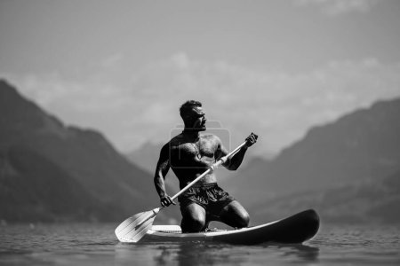 Foto de Male fit muscular model with paddle board. Guy paddling on paddleboard. Healthy summer lifestyle. Water sport. SUP surfing in summer vacation in Alps lake in Switzerland - Imagen libre de derechos