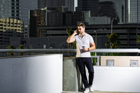 Portrait of handsome male model chatting on phone outdoor. Stylish man talking on phone dressed in polo. Fashion male posing on the street background. Business phone conversation. Urban style
