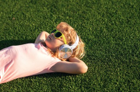 Cute and happy child 7-8 years old lying on the grass and listening to music in headphones on a sunny summer day