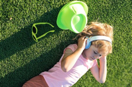 Emotional funny child in t-shirt in headphones in summer park. Children music concept. Little child wears stereo headphones, enjoys sound and listens favourite song