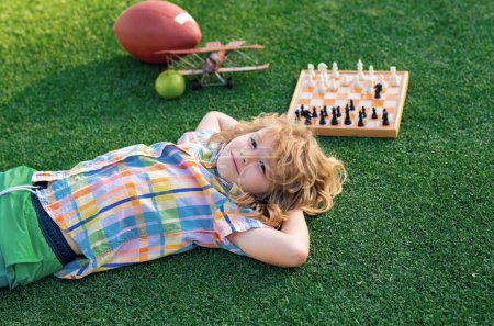 Chess game for children. Kid relax in park, laying on grass, daydreaming. Kid playing chess. Games and activities for children