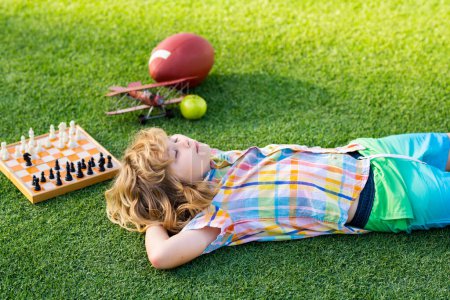 Kid relax in park, laying on grass, daydreaming. Little kid play chess in park. Child boy playing board game outdoor. Thinking child brainstorming and idea in chess game