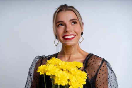 Photo for A beautiful woman in an elegant black dress with stunning evening makeup poses against a white studio backdrop with a bouquet of yellow flowers, exuding a festive and playful mood - Royalty Free Image