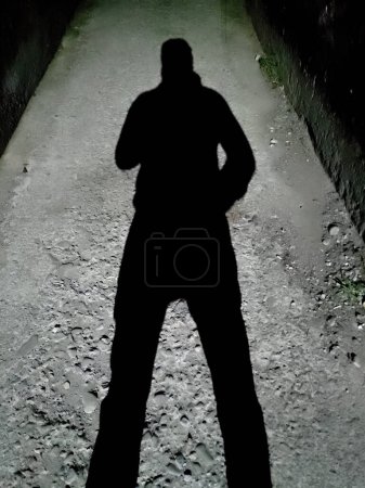 Hombre proyectando una sombra contra un poste de luz en un callejón oscuro por la noche. India.