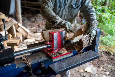 Caucasian senior man splits wood with a hydraulic log splitter. He wears a denim work shirt, work gloves and ear protection. Defocused wood pile in background, bark texture, wood splits in his hand.