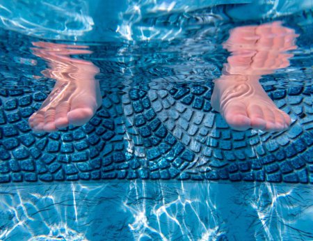 Foto de Lindo primer plano de los pies de los niños en una piscina azul idílico con fondo de azulejo azul decorativo y la luz del sol que se refleja en el lado. Ripplinf agua arriba con espacio para copiar. - Imagen libre de derechos