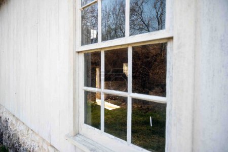 Téléchargez les photos : Vieille fenêtre reflète la nature dans un bâtiment colonial américain en bois blanchi à la chaux. Mur extérieur, intérieur de la pièce visible par la fenêtre. - en image libre de droit