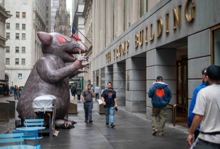 Foto de Nueva York, NY, Estados Unidos 10-06-2023 Los peatones se reúnen en la acera y la calle empedrada frente al edificio Trump en el distrito financiero de la ciudad de Nueva York. Una rata inflable gigante está delante de la puerta - Imagen libre de derechos