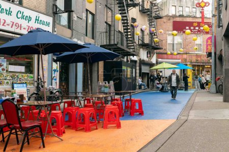 Téléchargez les photos : Chinatown, New York City, NY, États-Unis 11-09-2023 Des sièges en plastique rouge vif entourent les tables en couleurs arc-en-ciel peintes dans la rue Chinatown. Piétons et travailleurs, parasols et bâtiments colorés - en image libre de droit