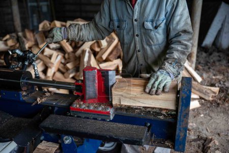 Photo for Caucasian senior man splits wood with a hydraulic log splitter. He wears a denim work shirt, work gloves. Defocused wood pile in background, splitter blade cuts into wood. - Royalty Free Image