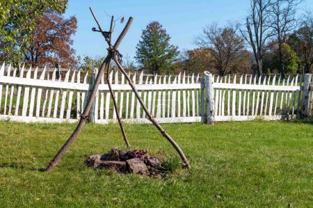 A white picket fence in the background and a colonial American backyard with a cook fire with tripod for hanging a cooking pot. Sunny day, no people, with copy space.