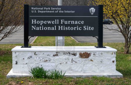 Foto de Hopewell Furnace National Park, Pensilvania, Estados Unidos, 13-03-2024. Señal de entrada con hierba y flores florecientes. Departamento del Interior de Estados Unidos, Servicio de Parques Nacionales. - Imagen libre de derechos
