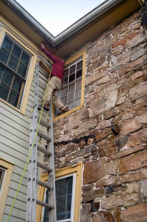 Niedriger Winkel Ansicht des Menschen auf Leiter Reparatur Dach des alten Steinhauses mit antiken Fenstern und bunten Feldstein. Architektonische Besonderheiten sichtbar.