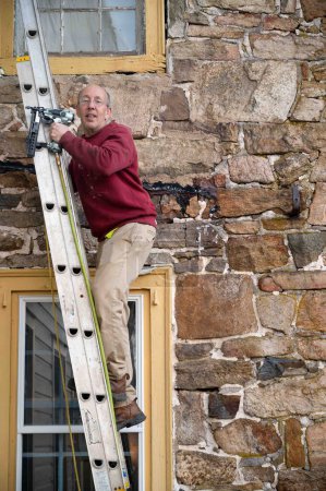 Téléchargez les photos : L'homme grimpe une échelle adossée à une ancienne ferme en pierre de taille avec des fenêtres antiques. Il porte un pistolet pneumatique dans sa main - en image libre de droit