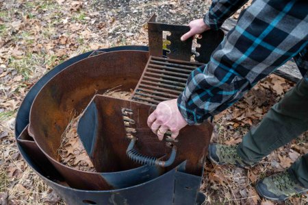 Man in a flannel shirt and hiking boots adjusts the grates on a campground fire pit grill. Autumn leaves on forest floor.. With some copy space
