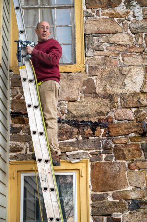 Hoch oben auf einer Leiter lehnt ein Mann an einem alten Bauernhaus mit antiken Fenstern. Er trägt eine Druckluftpistole in der Hand