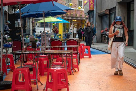 Téléchargez les photos : New York, NY, USA 11-09-2023 Tables de restaurant Chinatown vides sur Doyers Street coloré avec des chaises en plastique rouge et des piétons qui passent. Usage rédactionnel uniquement - en image libre de droit