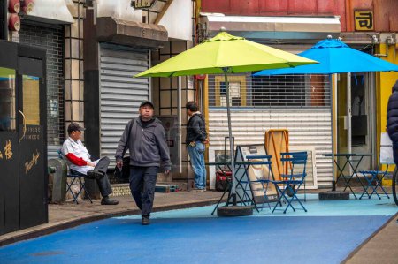 Téléchargez les photos : New York, NY, États-Unis 11-9-2023 Tables de reataurant extérieur vides sur Doyers Street à Chinatown avec parasols colorés. Travailleur dans le tablier sur pause à l'extérieur dans la chaise. Piétons et vitrines fermées. Usage rédactionnel uniquement. - en image libre de droit