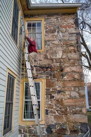 Téléchargez les photos : Un homme en bottes de travail monte une échelle adossée à une maison de ferme historique en pierre colorée de Pennsylvanie - en image libre de droit