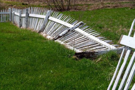 White handmade white picket fence with rotten fence post lays broken on the ground of lush green grass. No people, natural sunlight, copy space.