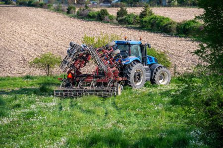 Primer plano de un tractor de granja azul tirando de un gran cultivador de gradas de disco implementar a través de la hierba verde en el campo agrícola. Luz del sol, sin gente, espacio para copiar.