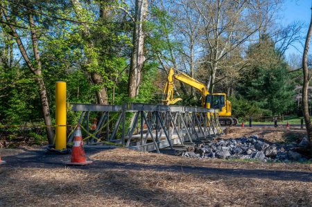 An excavator intalls new metal pedestrian foot bridge at public park with caution tape orance safety cone and stones