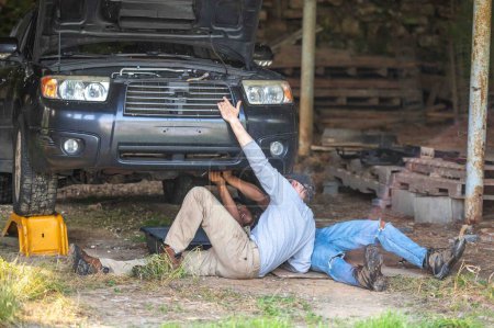 Père et fils au sol dans une grange rurale travaillant sur une voiture. La voiture a capot ouvert, jeune homme tourne clé. Famille interraciale.