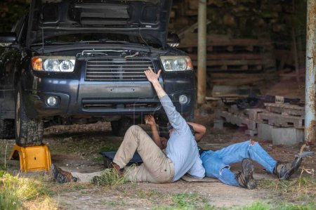 Father and son on the ground in a rural barn working on a car. Car has hood open, young man turns wrench. Interracial family.