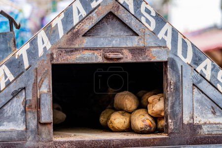 Téléchargez les photos : Four à rôtir les pommes de terre avec la phrase patatas asadas écrite en espagnol, travaillant au milieu d'un marché de rue médiéval. - en image libre de droit