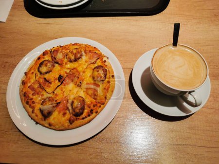Small pizza next to a huge XXL cup of coffee with milk and a spoon, on a wooden table.