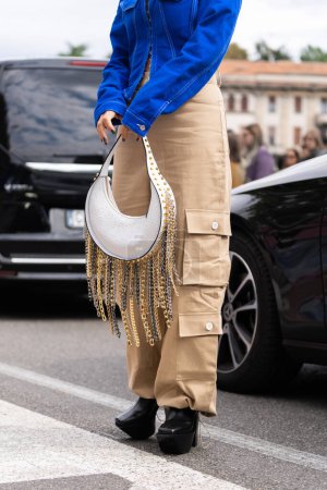 Foto de Milán, Italia - 21 de septiembre de 2022: mujer en chaqueta de mezclilla azul asimétrica recortada, bolso de cuero con clavado tachonado cadena flecos colgante hombro, pantalones acampanados de carga beige, plataforma zapatos de tacón alto. - Imagen libre de derechos