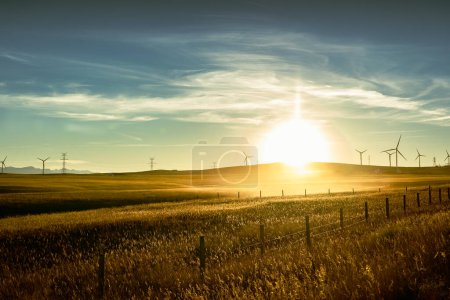 Foto de Salida del sol con niebla de la mañana sobre campos agrícolas cosechados con una cerca de alambre de púas y molinos de viento distantes que producen electricidad en un paisaje de pradera. - Imagen libre de derechos