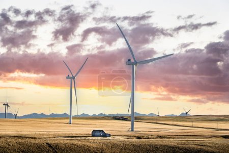 Sonnenuntergang Windmühlen produzieren grüne Energie mit Blick auf abgeerntete Felder und entfernte Berge mit einer rustikalen Scheune auf der kanadischen Prärie.
