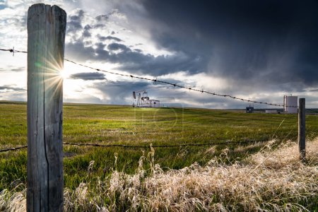 Photo for Prairie landscape scene with barbed wire fence overlooking a distant oil pump jack under a stormy sky in Rocky View County Alberta Canada. - Royalty Free Image