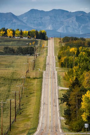 Photo for Rural road lined with telephone poles and autumn trees in fall colours with Canadian Rocky Mountains at background in Rocky View County Alberta Canada. - Royalty Free Image