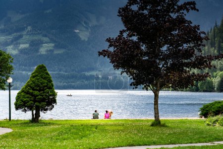 Photo for Couple sitting down on a shoreline overlooking a lake with landscaped trees and mountains at background enjoying summer vacation in British Columbia Canada. - Royalty Free Image