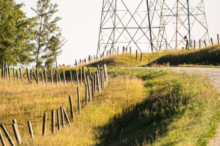 Wooden fence posts line the curve or a rural gravel road overlooking a distant transmission tower in Rocky View County Alberta Canada.