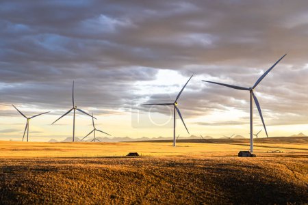 Reihe von Windmühlen, die auf der kanadischen Prärie Strom erzeugen, mit Blick auf ein Paar in alten Bauernschuppen unter einem dramatischen Sonnenuntergang in der Nähe von Pincher Creek Alberta.