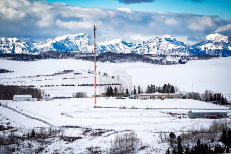 Oil and gas industry flare stack overlooking snow covered hills with Alberta Rocky Mountains at background in Western Canada.