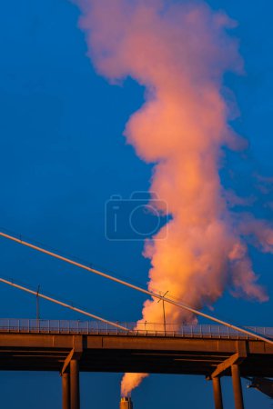 Photo for Industrial chimney by bridge billowing smoke into blue sky, low angle view, vertical image, Gothenburg, Sweden, Europe - Royalty Free Image