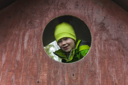 Photo for A young boy wearing a green hat and jacket looks out of a red house, playing in the playground. - Royalty Free Image